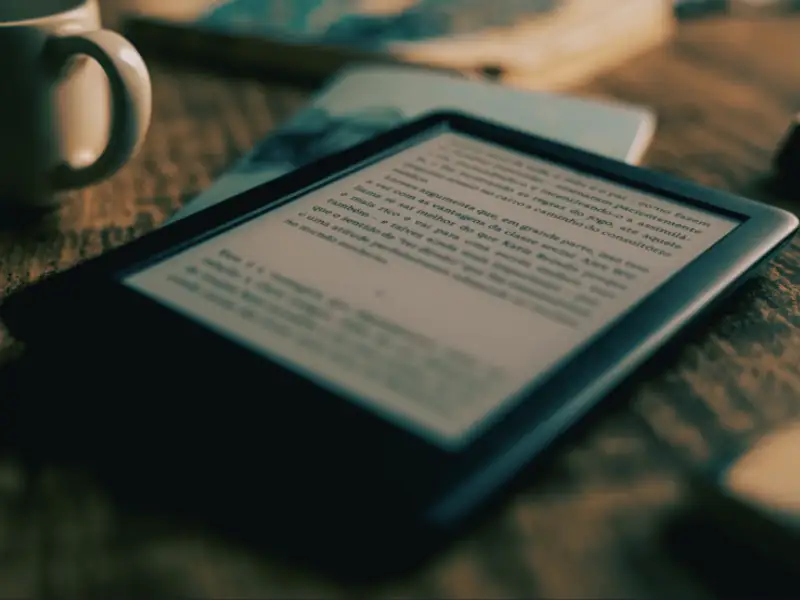 Un kindle sobre la mesa con un libro electrónico abierto, un café y un libro físico debajo.